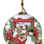 Hand-painted glass ornament featuring a snowman family sitting in the back of a red truck, surrounded by trees and presents in a snowy holiday scene.
