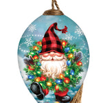 Hand-painted glass ornament featuring a festive gnome holding a wreath decorated with colorful Christmas lights against a snowy backdrop.